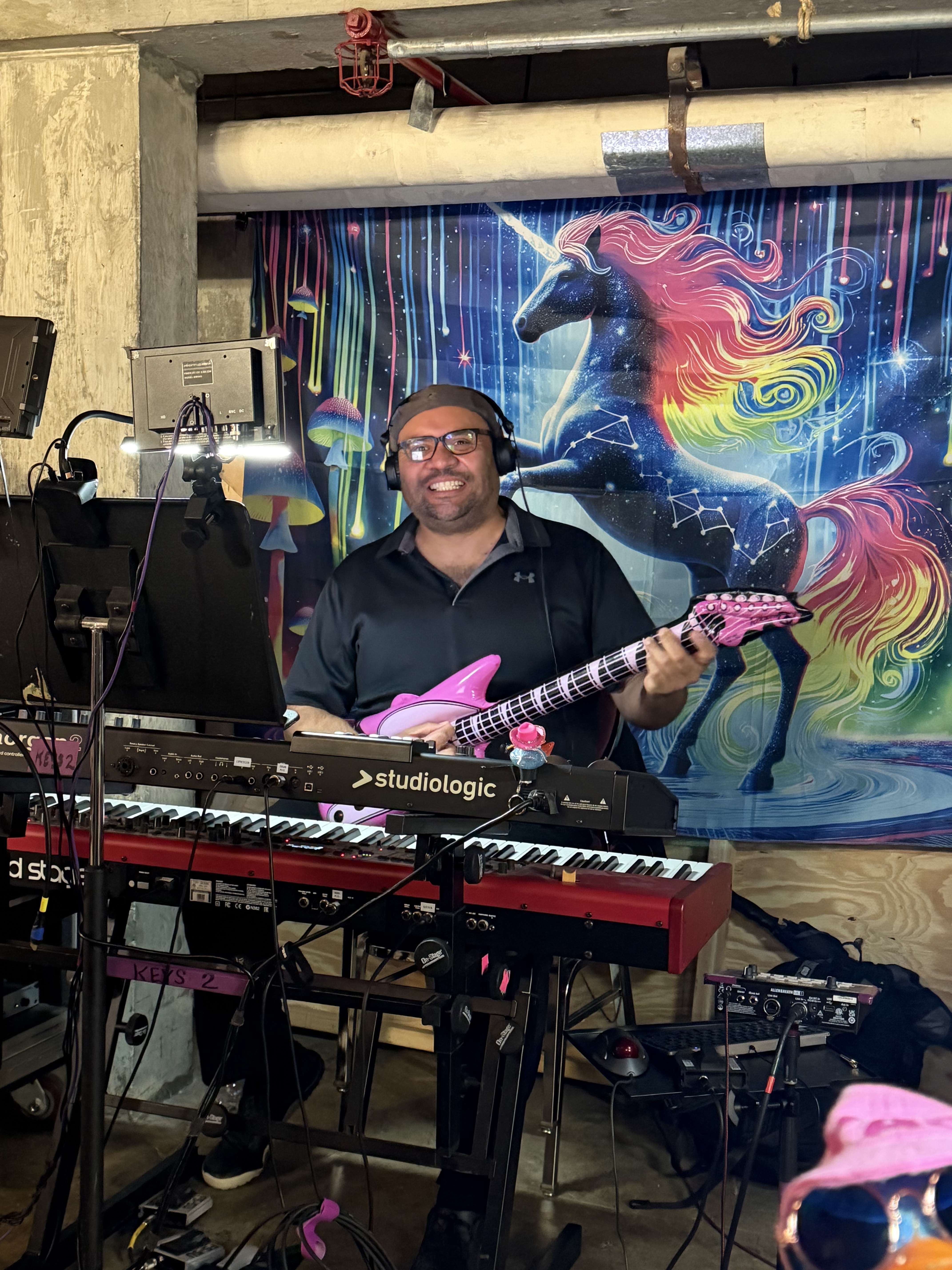 Nathan Spear smiling with a pink guitar behind keyboards, a rainbow unicorn backdrop behind him.