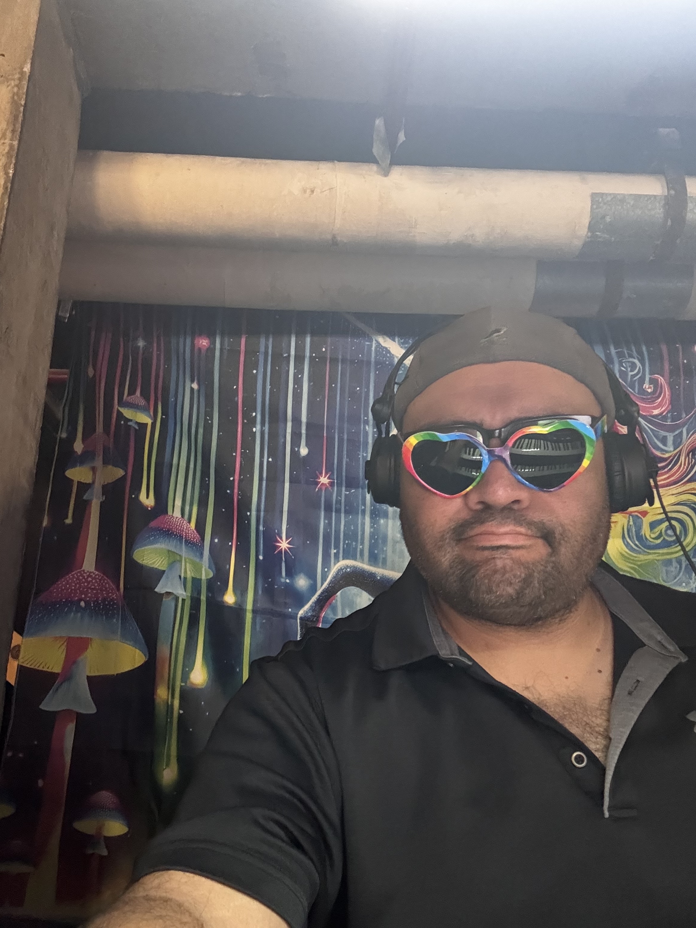 Nathan Spear in rainbow heart sunglasses and headphones in front of a psychedelic mushroom mural.